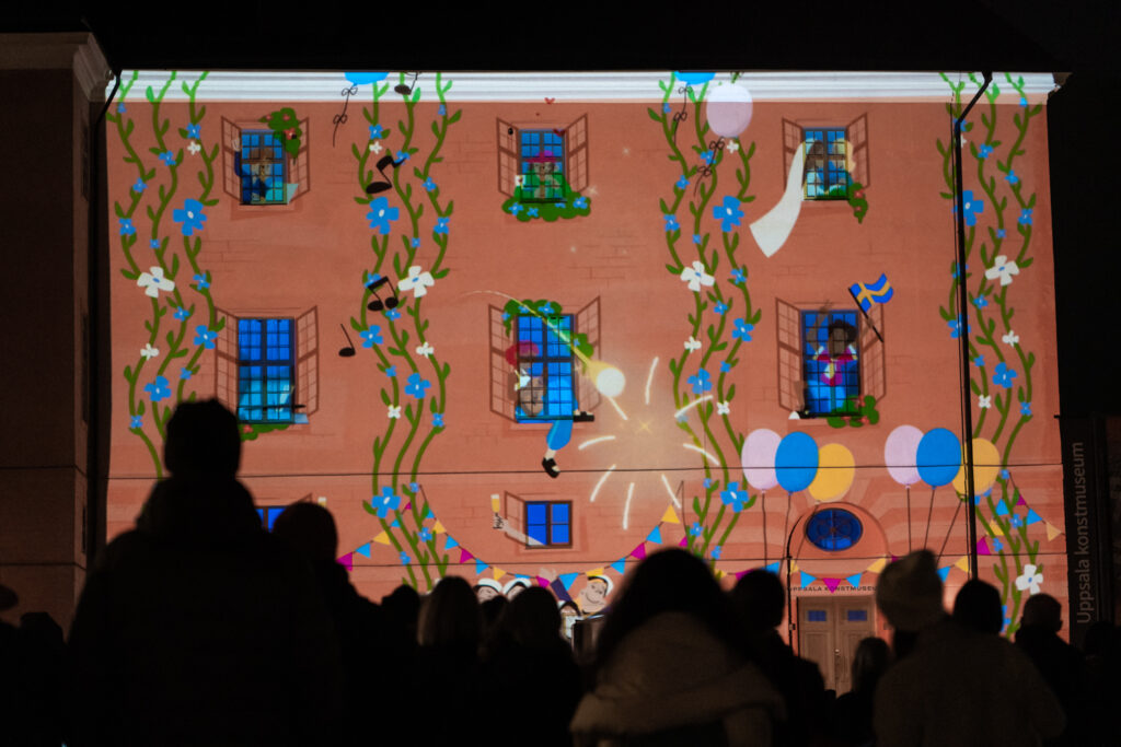 Tecknade singerväxter, fönster, flagga och musiknoter projicerade på en slottsfasad. Människor i förgrunden.